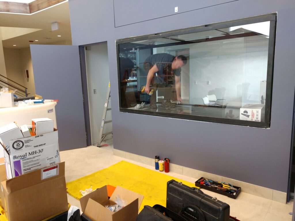 Two workers install a large glass window in an interior wall; tools and boxes are scattered on the floor nearby.