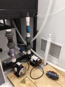Two electric water pumps connected to PVC piping are mounted on a wooden platform near a vent in a utility room.