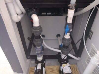 Plumbing system with pipes, valves, and pumps under a tank, installed on a wooden platform in a utility area.