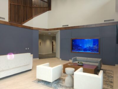 Modern lobby with white chairs, a gray couch, a wooden table, and a built-in aquarium on a blue accent wall.
