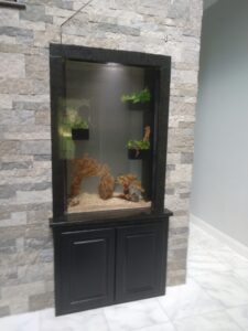 A built-in aquarium with plants and rocks is set into a stone wall above black cabinet doors on a marble floor.