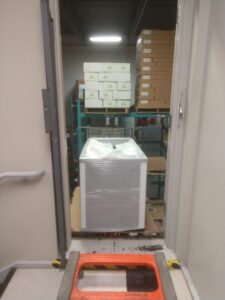 Pallets of boxed items are stacked in a storage room with metal shelving and an open door in the foreground.