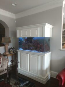 Large aquarium with rocks and sand sits in a white cabinet against a beige wall in a living room setting.