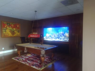 A pool table with a Budweiser light, a colorful rug, a wall aquarium, and a painting in a wood-paneled room.