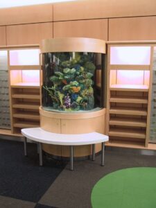 A cylindrical aquarium is set in a wooden display with empty shelves and a white curved bench in front.